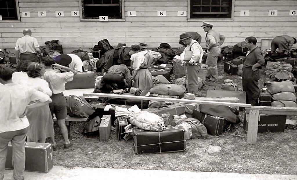 Foreground: Refugees with luggage; Background: Barrack-homes with letters on exterior to organize the refugees